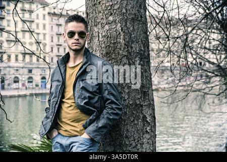 Three-quarter length of contemplative light brown haired young man wearing grey jacket and denim jeans standing beside picturesque river in Turin, Ita Stock Photo