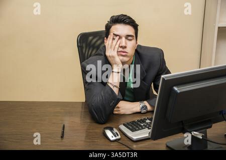 Tired bored young businessman sitting at his desk in front of his computer with his chin resting on his hand and eyes closed Stock Photo
