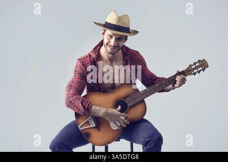 Studio portrait of handsome bearded man with unbuttoned shirt playing guitar over grey background, sitting on chair, looking at camera Stock Photo
