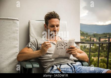 Young man sitting doing a crossword puzzle looking thoughtfully at a magazine, as he tries to think of the answer to the clue Stock Photo