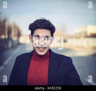 Handsome young man in city street, looking at camera smiling, wearing suit jacket and turtleneck Stock Photo