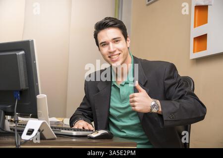 Smiling handsome young businessman sitting at his desk in the office looking at camera, doing thumb up sign Stock Photo