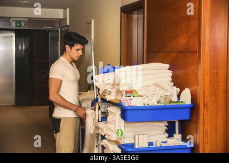 Young man pushing a housekeeping cart laden with clean towels, laundry and cleaning equipment in a hotel as he services the rooms Stock Photo