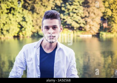 Portrait of contemplative light brown haired young man wearing white shirt beside picturesque river or lake, looking away to a side during fall Stock Photo