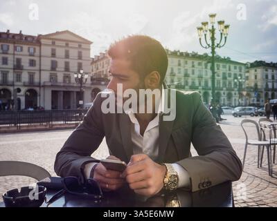 Handsome young man using cellphone, wearing elegant coat posing at bar table outside on urban background Stock Photo