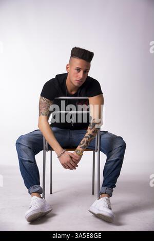 Trendy tattooed young man sitting on chair backwards, on light background Stock Photo