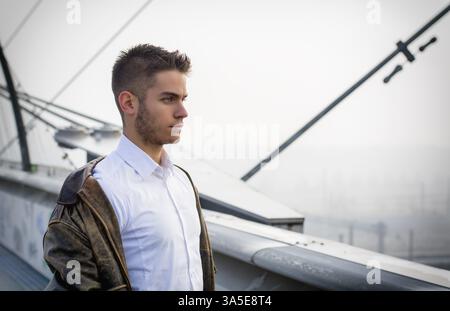 Handsome trendy young man standing on a sidewalk wearing a fashionable jacket and scarf in a relaxed confident pose looking away to a side Stock Photo