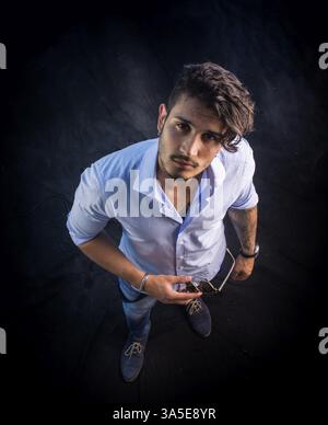 Portrait of brunette young man in light blue shirt and jeans, standing in studio shot from above perspective, against dark background. Full length pho Stock Photo
