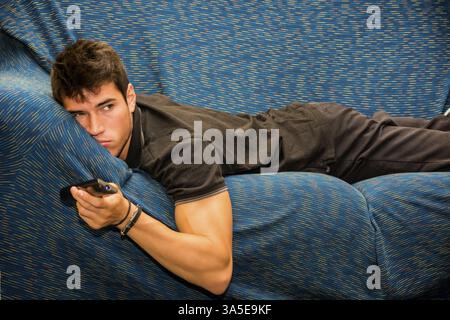 Young bored man sitting on sofa watching television changing the channel with the remote control Stock Photo