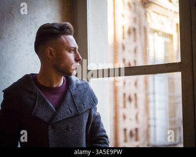 Handsome young serious man standing inside old building next to big window, wearing wool sweater, looking out Stock Photo