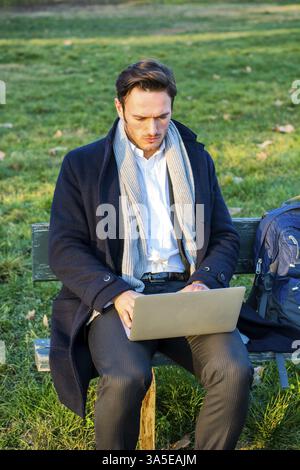 Handsome elegant businessman sitting on a wooden bench working outdoors in an urban park typing information onto his laptop computer with a serious ex Stock Photo