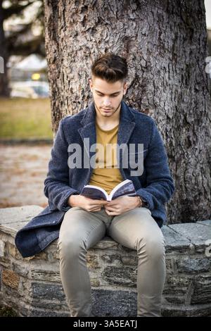 Male student reading book outdoor. Handsome young man reads a book sitting outside on a bench in city park Stock Photo