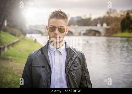 Three-quarter length of contemplative light brown haired young man wearing grey jacket and denim jeans standing beside picturesque river in Turin, Ita Stock Photo