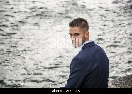 Three-quarter length of contemplative light brown haired young man wearing grey jacket and denim jeans standing beside picturesque river in Turin, Ita Stock Photo