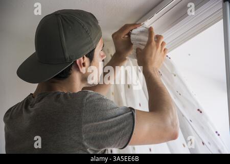 Young handsome man or handyman installing curtains over window at home, doing renovation work Stock Photo