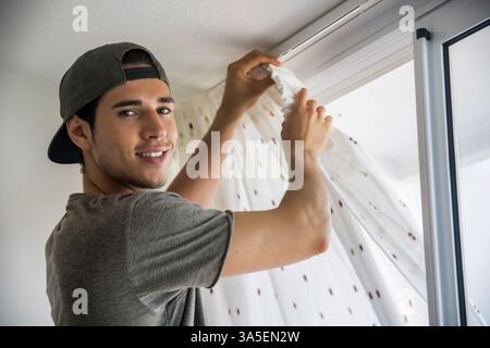 Young handsome man or handyman installing curtains over window at home, doing renovation work Stock Photo