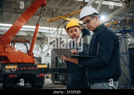Architect is showing the plan on the laptop. Two male factory workers are in the warehouse. Stock Photo