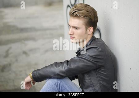 Attractive blond young man wearing black leather jacket and jeans sitting on stairs outside Stock Photo