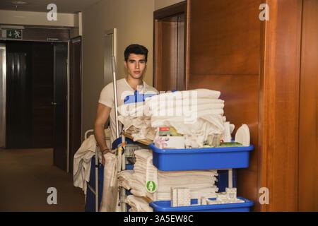 Young man pushing a housekeeping cart laden with clean towels, laundry and cleaning equipment in a hotel as he services the rooms Stock Photo