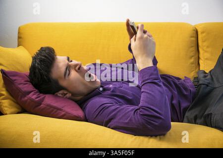 Attractive young man using cell phone and yawning, tired, while laying on couch, with serious expression Stock Photo