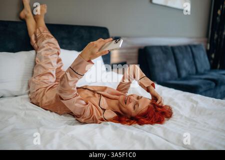 Relaxing, lying down on bed with smartphone in hands. Woman is in the hotel room, modern interior. Stock Photo