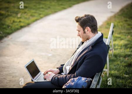 Handsome elegant businessman sitting on a wooden bench working outdoors in an urban park typing information onto his laptop computer with a serious ex Stock Photo
