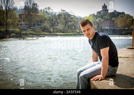 Three-quarter length of contemplative light brown haired young man wearing dark t-shirt and denim jeans sitting on wall beside picturesque river in Tu Stock Photo
