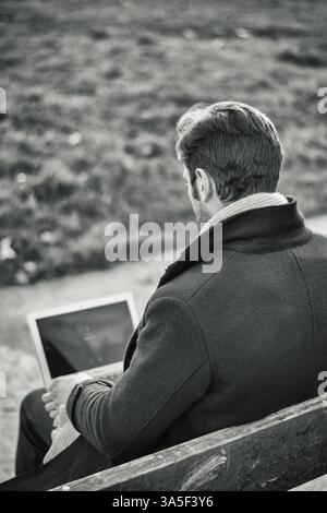 Handsome elegant businessman sitting on a wooden bench working outdoors in an urban park typing information onto his laptop computer, seen from the ba Stock Photo