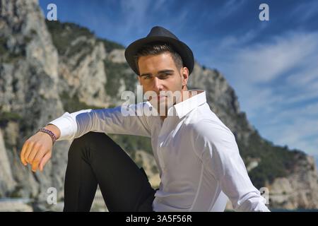 Young handsome man in elegant white shirt and black fedora hat, outdoor while looking at camera. Bright blue sky behind Stock Photo