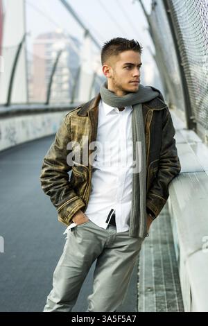 Handsome trendy young man standing on a sidewalk wearing a fashionable jacket and scarf in a relaxed confident pose looking away to a side Stock Photo