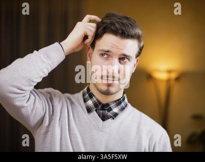 Confused or doubtful young man scratching his head and looking up. Indoors shot in a living room Stock Photo