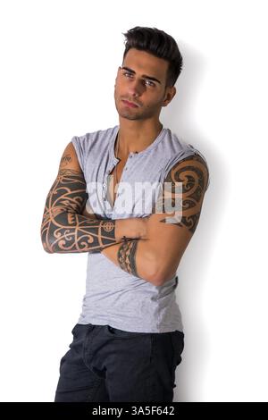 Handsome tattooed young man wearing grey t-shirt, standing against light background in studio shot with arms crossed on chest Stock Photo