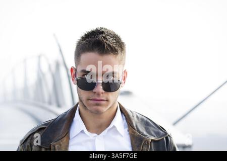 Handsome trendy young man standing on a sidewalk, wearing sunglasses and a fashionable leather jacket in a relaxed confident pose looking at camera ov Stock Photo