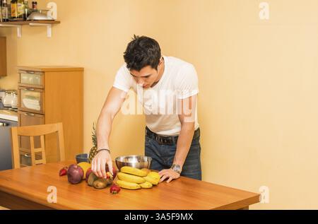 Young man preparing a healthy fruit salad or smoothie, standing leaning on a kitchen counter loaded with an assortment of fresh tropical fruit, lookin Stock Photo