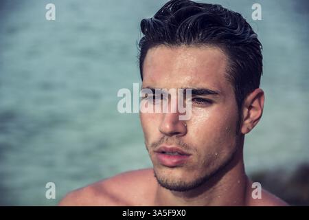 Attractive young man in the sea getting out of water with wet hair, looking away to a side Stock Photo