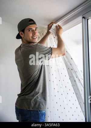 Young handsome man or handyman installing curtains over window at home, doing renovation work Stock Photo