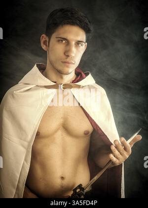 Handsome young man in old fashioned cloak with sword on dark smoky background Stock Photo
