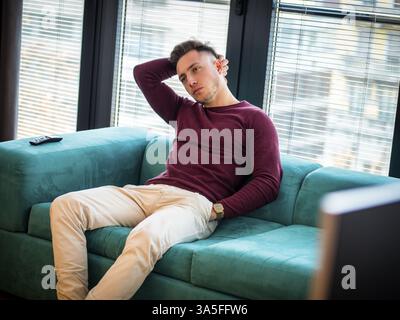 Young bored man lying on sofa watching television changing the channel with the remote control Stock Photo