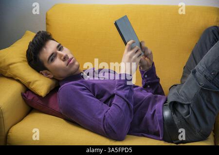 Attractive young man using tablet PC while laying on couch, looking serious at camera Stock Photo