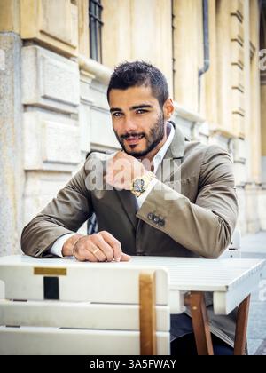 Handsome young man wearing elegant coat posing at table outside on urban background Stock Photo