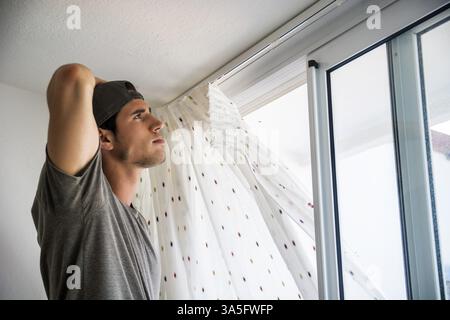 Young handsome man or handyman installing curtains over window at home, doing renovation work Stock Photo