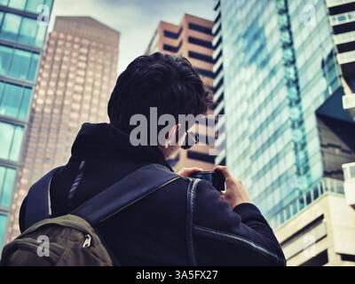 Young man taking photograph with smartphone in modern city, in a winter or fall day, seen from the back Stock Photo