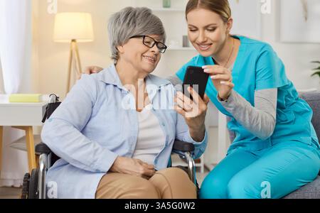 Smiling elderly woman in wheelchair using smartphone while sharing something funny with nurse. Stock Photo