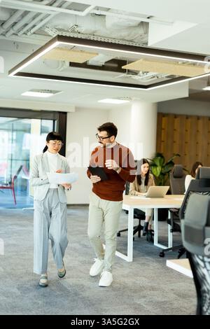 Two colleagues engage in a dynamic discussion while walking through a contemporary office filled with creativity. Others focus on their tasks, showcas Stock Photo