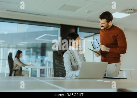 Two colleagues engage in a lively discussion in a contemporary office, exchanging ideas and insights while another team member works focused in the ba Stock Photo