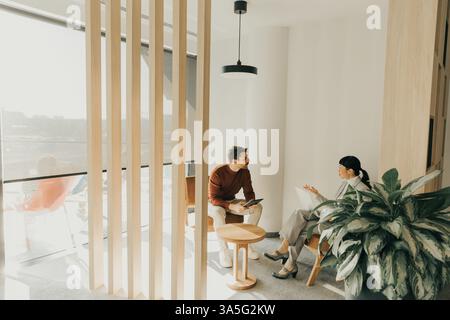Two individuals in contemporary attire converse intently in a bright lounge featuring wooden accents and greenery. The sunlight casts soft shadows, ad Stock Photo