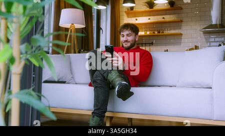 Happy Young Man Using Smartphone While Relaxing on Couch in Cozy Home, Smiling and Engaged in Online Communication Stock Photo