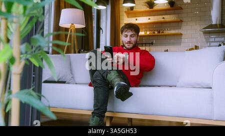 Young Man Using Smartphone While Relaxing on Couch in Cozy Home, Smiling and Engaged in Online Communication Stock Photo