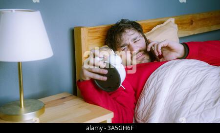 Young man in bed answering phone call from work, holding alarm clock, unhappy about waking up early, stressed morning routine Stock Photo