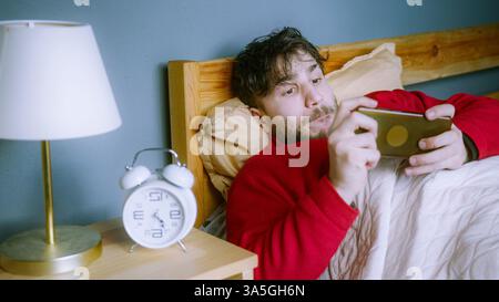 Young man in red sweater lying in bed, playing mobile game with intense focus, ignoring alarm clock in cozy bedroom, morning routine concept Stock Photo